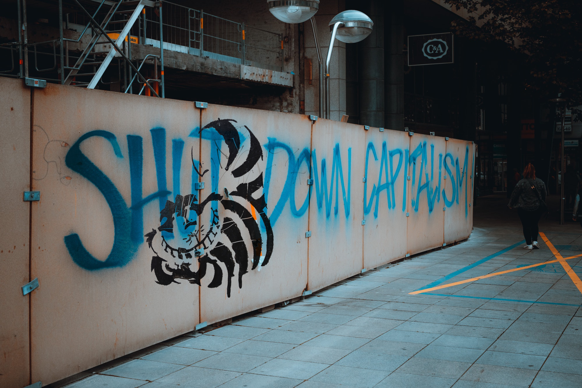 Graffiti of the Cheshire cat from Alice in Wonderland superimposed (in Photoshop) over an image of graffiti reading "Shut Down Capitalism" on a wall, with a figure walking by