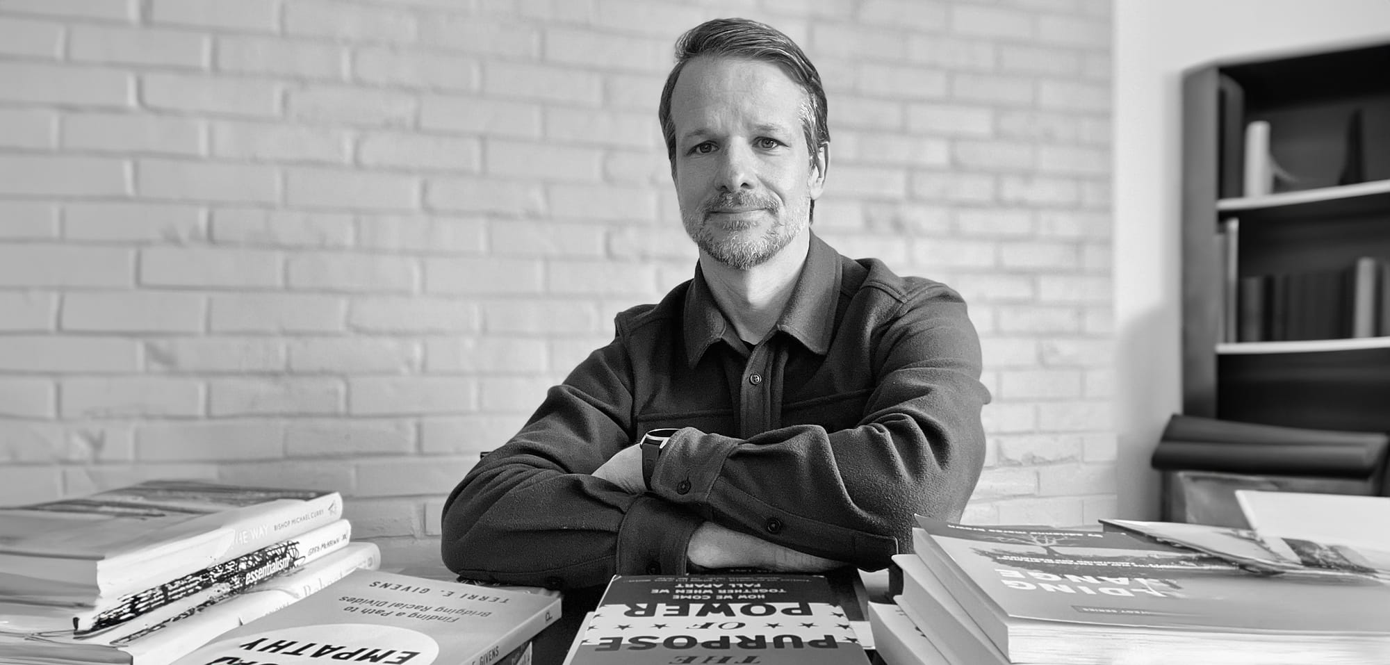 a black-and-white photo of a man with a light beard sitting at a table, facing the viewer, surrounded by stacks of books