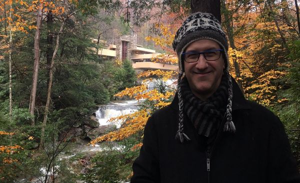 a man in a hat and glasses in front of a modernist home (Fallingwater) above a flowing stream