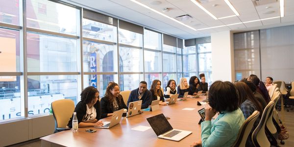 about twenty people, all with laptops or tablets, meeting at a large table in a room with a large window